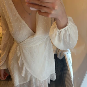 Vit omlottklänning med volang - Säljer en superfin vit omlottklänning med volangdetaljer och långa ärmar. Klänningen har en lätt transparent känsla och knyts i midjan för en snygg passform. Perfekt för sommarens alla tillfällen!