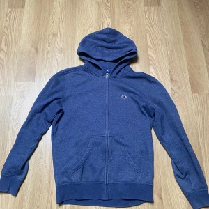 Blå hoodie från Champion - Säljer en blå hoodie från Champion med dragkedja framtill och huva. Tröjan har långa ärmar och en klassisk känguruficka. Liten Champion-logga på bröstet. Perfekt för en avslappnad stil.