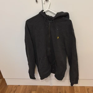 Mörkgrå hoodie från Lyle & Scott - Säljer en mörkgrå hoodie från Lyle & Scott med dragkedja och huva. Tröjan har ett broderat gult fågelmärke på bröstet och två fickor framtill. Perfekt för en avslappnad stil.
