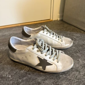 Golden Goose  - Säljer ett par vita Golden Goose Super-Star sneakers med grå stjärna på sidan och grå häl. Skorna har grå snörning och klassisk låg modell. Perfekta för dig som gillar en stilren men ändå unik look, box ingår och tveka inte på att fråga om ni funderar över något!