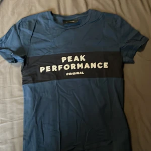 Blå t-shirt från Peak Performance - Säljer en blå t-shirt från Peak Performance med vit logga och text på bröstet. T-shirten har en klassisk passform och är kortärmad, perfekt för vardagsbruk eller träning.