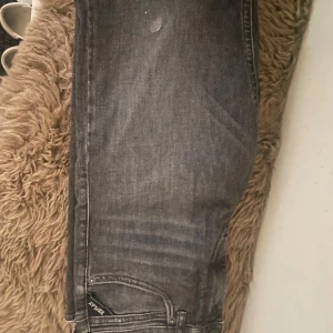Grå jeans från Replay - Säljer ett par grå jeans från Replay med klassisk femficksdesign och raka ben. Jeansen har en diskret tvättad look och tydlig Replay-lapp bak i midjan. Perfekta för dig som gillar en enkel och stilren stil. Den har dock en liten fläck på benet