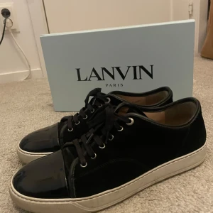 Svarta sneakers från Lanvin - Säljer ett par svarta sneakers från Lanvin med vit platt sula och rund tå. Skorna har snörning och är tillverkade i blankt skinn. Klassisk och stilren design med diskreta detaljer. Skokartong och dustbags medföljer.