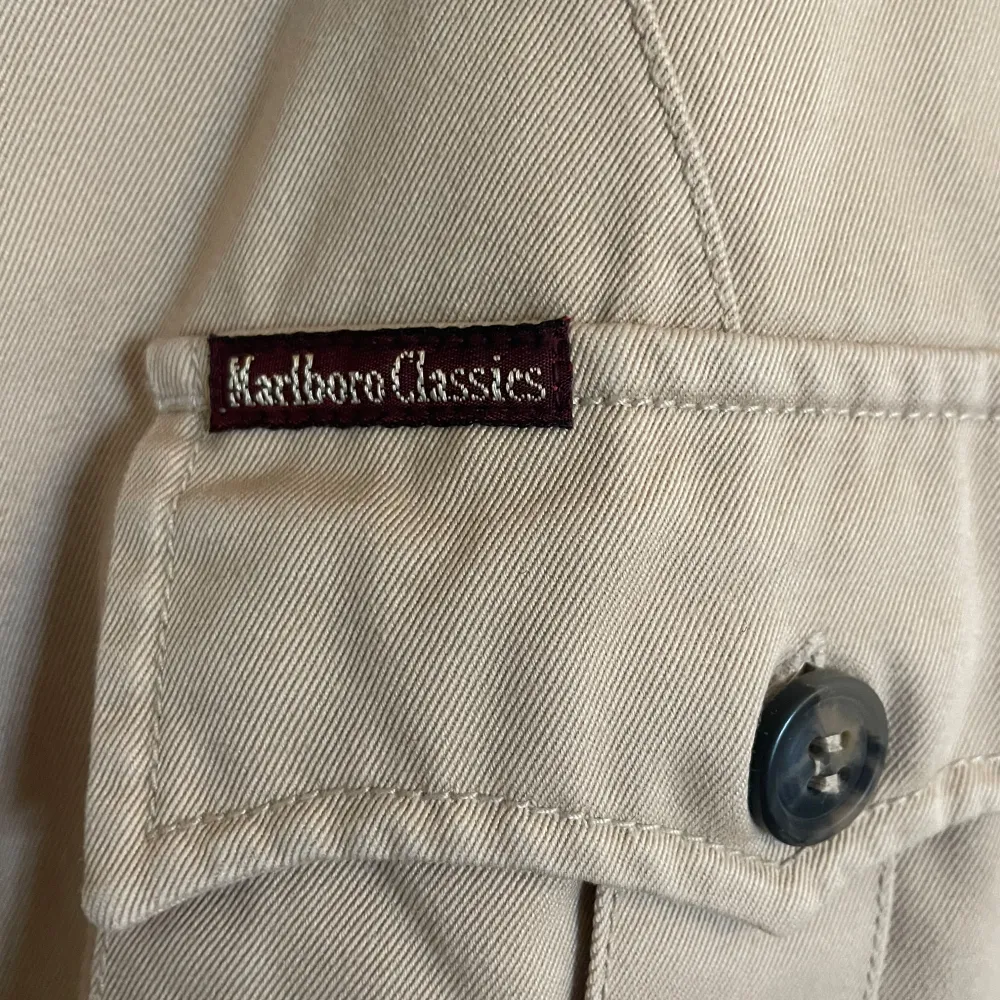 Klassisk beige utilityjacka från Marlboro Classics med fyra stora fickor framtill och knäppning med knappar. Jackan har en avslappnad passform och tydlig krage. Perfekt för dig som gillar tidlös och funktionell stil.. Takit.