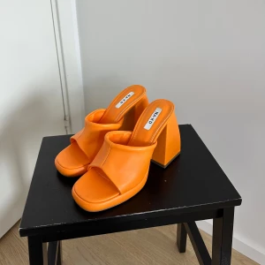 Orange klackade sandaler från NA-KD  - Säljer ett par trendiga orange sandaler från NA-KD med bred rem och öppen tå. Skorna har en hög, kraftig klack och platt sula framtill. Perfekta för att addera färg till din outfit!