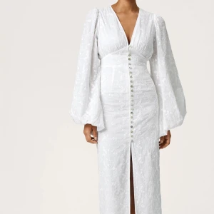 Vit långklänning Positano från Adoore - Superfin vit långklänning från Adoore i modellen Positano. Klänningen har v-ringning, långa puffärmar och knappar framtill. Tunt blommigt mönster i tyget och slits framtill. Perfekt för sommarens speciella tillfällen! Endast provad