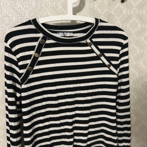 Randig ribbad tröja med knappar från stradivarius  - Säljer en svartvit randig ribbad tröja från stradivarius med dekorativa knappar längs axlarna. Tröjan har lång ärm och rund halsringning. Perfekt för en stilren och trendig look.