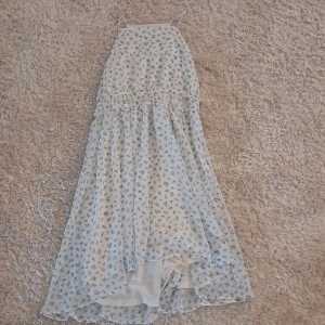 Vit blommig sommarklänning från H&M - Söt vit klänning från H&M med små blå blommor och smala axelband. Klänningen har en luftig passform och är tillverkad i ett lätt, flowy material. Perfekt för varma dagar och har en något längre bakdel för extra stil.