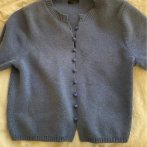 Blå kofta med klädda knappar - Säljer en ljusblå kofta med rund halsringning och korta ärmar. Koftan har klädda knappar framtill och ribbade muddar vid ärmslut och nederkant. Perfekt att slänga över en topp för en chill look.