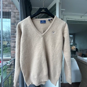 Beige v-ringad ulltröja från Gant - Säljer en beige v-ringad tröja från Gant i superfin lammull. Tröjan har långa ärmar och ribbade muddar vid ärmslut och nederkant. Perfekt för lager-på-lager och en stilren look. Den är lite liten i storleken.
