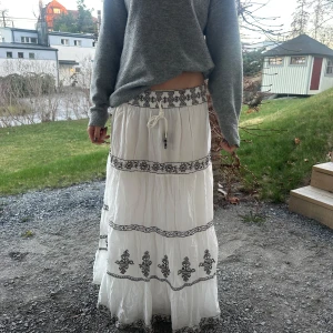 Lång kjol  - Säljer en vit långkjol från Zara med svarta broderade mönster och knytband i midjan. Kjolen har flera våder med dekorativa broderier och en luftig, bohemisk känsla. 