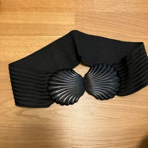 Svart brett midjebälte med snäckformade detaljer - Säljer ett svart elastiskt midjebälte med två stora snäckformade spännen framtill i metall-look. Bältet är brett och ger en markerad midja, perfekt för att lyfta en outfit med en cool detalj.