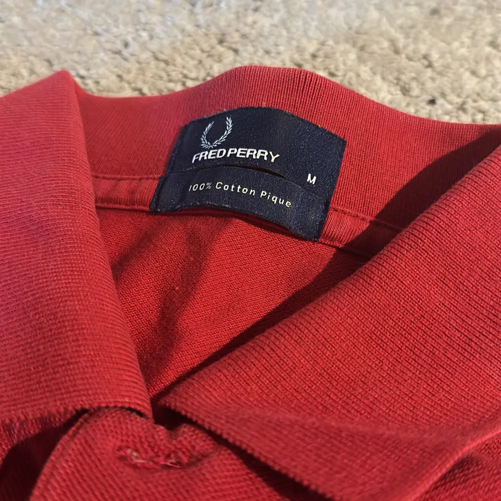 Säljer en klassisk röd piké t-shirt från Fred Perry med vit broderad logga på bröstet. T-shirten har krage, knappar vid halsen och är tillverkad i 100% bomull. Perfekt för en stilren och sportig look.. T-paidat.