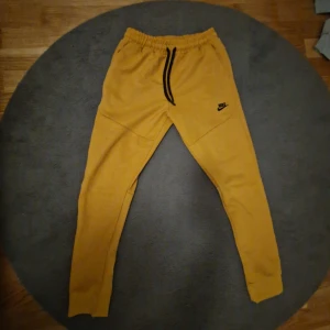 Gula mjukisbyxor Nike tech  - Säljer ett par gula mjukisbyxor från Nike med svart dragsko i midjan och logga på vänster ben. Byxorna har en slim passform och är perfekta för träning eller chill. Mjukt bomullsmaterial och elastisk midja för extra komfort.