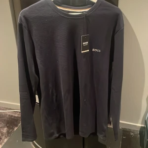 Mörkblå långärmad tröja från BOSS - Säljer en stilren mörkblå långärmad tröja från BOSS med diskret logga på bröstet. Tröjan har rund halsringning och är tillverkad i ett mjukt, ribbat material som ger en snygg struktur. Perfekt för en clean och klassisk look.