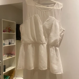 Vit ärmlös blus från Vero Moda - Säljer en vit ärmlös blus från Vero Moda med volangdetaljer och lätt transparent tyg. Blusen har en luftig passform och en öppen detalj i ryggen. Perfekt för varma dagar eller att styla lager på lager.