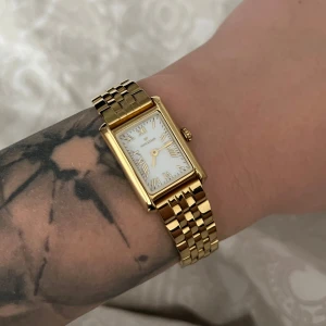 Guldfärgad klocka från Mockberg - Säljer en guldpläterad klocka från Mockberg med rektangulär vit urtavla och romerska siffror. Klockan har en klassisk länkarmband i rostfritt stål och levereras i originalask med extra länkar.