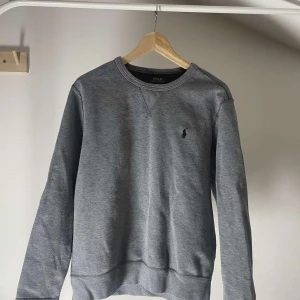 Grå sweatshirt från Polo Ralph Lauren - Klassisk grå sweatshirt från Polo Ralph Lauren med rund halsringning och broderad logga på bröstet. Tröjan har långa ärmar och ribbade muddar vid ärmslut och nederkant. Perfekt för en avslappnad stil.