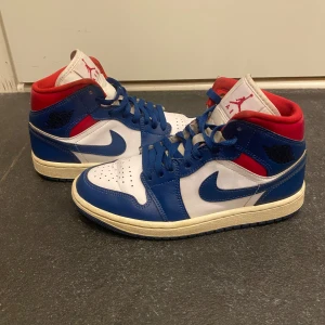 Nike Air Jordans blå, vita och röda - Säljer ett par Nike Air Jordans med blå, vita och röda paneler. Skorna har klassisk hög siluett, snörning och rund tå. Ovandelen är i skinn med perforerad tåbox och vit sula. Perfekta för dig som gillar streetwear och sneakerskultur.