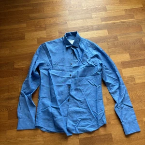 Blå skjorta i bomull - Snygg blå skjorta med klassisk krage och knäppning framtill. Skjortan har lång ärm och är tillverkad i ett mjukt bomullstyg. Perfekt för dig som gillar en stilren look.