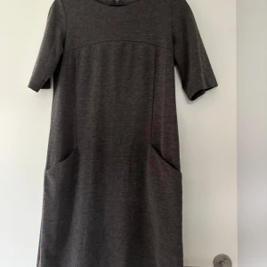 Mörkgrå klänning från Park Lane - Säljer en enkel och stilren mörkgrå klänning från Park Lane i storlek 38. Klänningen har korta ärmar, rund halsringning och praktiska fickor framtill. Passformen är normal och materialet känns mjukt och bekvämt.