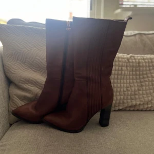 Bruna höga boots med klack och dragkedja - Säljer ett par bruna höga boots i skinn med markerad klack och dragkedja på insidan. Skorna har spetsig tå och dekorativa sömmar längs sidorna. Perfekta för dig som vill ha en stilren och trendig look.