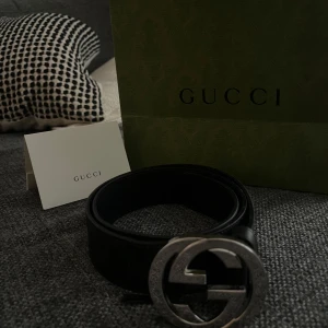 Svart bälte från Gucci med silverfärgat spänne - Säljer ett svart bälte från Gucci med ett stort, rundat spänne i silver med dubbla G-loggan. Bältet är i skinn och har diskret GG-mönster längs bandet. Perfekt accessoar för att lyfta din outfit.