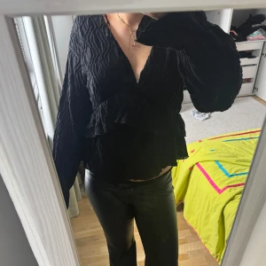 Svart blus med volang och v-ringning - Säljer en svart blus med v-ringning och volangdetaljer. Blusen har långa ärmar och en lätt skrynklig struktur i tyget. Perfekt för dig som gillar en stilren men ändå trendig look.