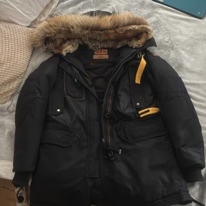 Svart parkajacka med pälskrage från Parajumpers - Säljer en svart parkajacka från Parajumpers med avtagbar pälskant på huvan och flera praktiska fickor. Jackan har gul detalj vid dragkedjan och är perfekt för kalla dagar. Jackan är ny och inte använd då den inte passar mig. Pris kan diskuteras!!