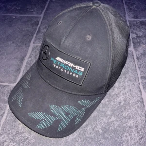 Svart Mercedes AMG Petronas keps - Snygg svart keps med Mercedes AMG Petronas Motorsport-logga framtill och dekorativa mönster i turkos på skärmen. Perfekt accessoar för motorsportfans.