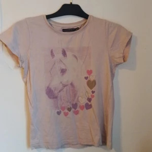 Ljusroa t-shirt med hästmotiv från Copperfield - Säljer en beige t-shirt från Copperfield med ett stort tryck av en häst och flera hjärtan i rosa, lila och beige på framsidan. T-shirten har korta ärmar och rund halsringning. Perfekt för dig som älskar hästar!