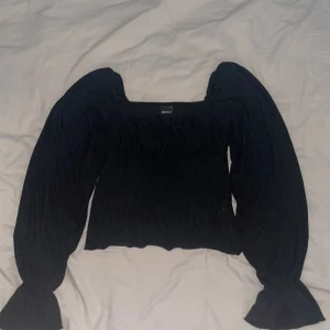 Svart långärmad blus med puffärmar - Säljer en svart blus med fyrkantig ringning och långa puffärmar. Toppen har en smockad kropp och vida ärmslut som ger en trendig siluett. Perfekt att styla med jeans eller kjol för en cool look.