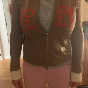 Brun ziptröja med röda bokstäver från Odd mollyMolly - Brun ziptröja från Odd Molly med stora röda bokstäver på framsidan. Tröjan har långa ärmar med beige muddar och dragkedja framtill. Perfekt för en avslappnad och trendig look.