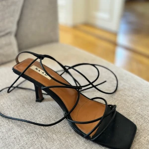 Svarta klackskor med snörning från NA-KD - Säljer ett par svarta sandaler från NA-KD med tunna remmar och snörning runt vristen. Har endast använt en gång så det är bara små slitningar undertill. Säljer då jag inte fått användning av dem.