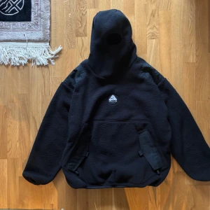 Svart Ninja fleece från Nike ACG - Säljer en svart fleecejacka från Nike ACG med stor huva och inbyggd ansiktsmask. Jackan har två fickor framtill med lock och dragkedja. Perfekt för kalla dagar och outdoor-äventyr. Klassisk ACG-logga på bröstet.