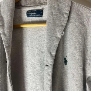 Grå cardigan jacka från Polo Ralph Lauren - Grå cardigan jacka från Polo Ralph Lauren med klassisk krage och broderad logga på bröstet. Jackan har två fickor framtill och är tillverkad i mjuk bomull. Perfekt för en avslappnad och stilren look.