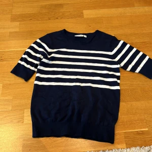 Marinblå och vit randig tröja från Kappahl - Säljer en marinblå tröja med vita horisontella ränder från Kappahl. Tröjan har rund halsringning och korta ärmar. Perfekt för en stilren och avslappnad look.