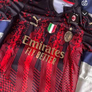 AC Milan fotbollströja Puma - Snygg AC Milan fotbollströja från Puma med digitalt röd-svart mönster och guldiga detaljer. Tryck med klubbmärke, sponsorloggor och texten 'SEMPRE MILAN' i nacken. Perfekt för fans som vill sticka ut på läktaren.