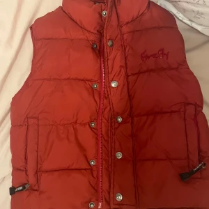 Röd dunväst från Firefly - Röd dunväst från Firefly med broderad logga på bröstet. Västen har både dragkedja och knappar framtill samt två fickor med detaljerade dragkedjor. Tillverkad i nylon och polyester, perfekt för lager-på-lager. storlek 130