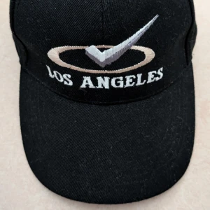 Svart keps med broderad Los Angeles-logga - Svart keps med böjd skärm och broderad text 'Los Angeles' framtill. Ovanför texten finns en broderad beige cirkel och en silverfärgad bock. Justerbar baktill för bra passform. Kepsen har aldrig använts 