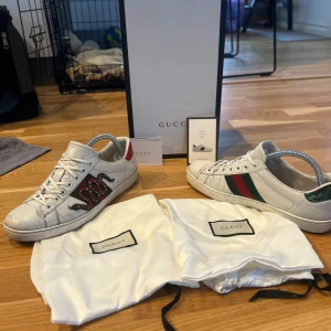 Gucci Ace - Gucci Ace sneakers i vitt skinn med klassiska gröna och röda ränder på sidan samt broderad orm. Baksidan har metallic-detaljer i rött och grönt. Skorna har rund tå och platt sula. Säljes med originalpåse och låda. Skorna är i 8/10. Box, dust bag, kort tillkommer✅hör av dig vid eventuella frågor 😊