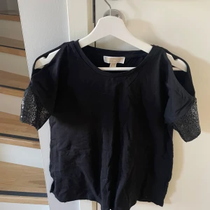 Svart topp från Michael Kors - Snygg svart topp från Michael Kors med unika detaljer. Toppen har korta ärmar med spets och öppna axlar som ger en trendig look. Perfekt för att lyfta vilken outfit som helst!
