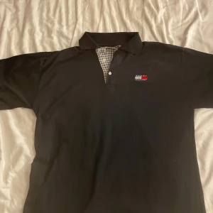 Svart pikétröja från Tommy Hilfiger - Snygg svart pikétröja från Tommy Hilfiger med klassisk krage och knappdetalj. Den har en liten logotyp på bröstet och ett rutigt mönster på insidan av kragen. Perfekt för en stilren look!