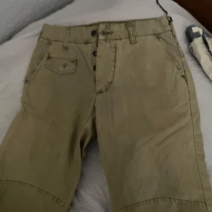 Beige bomullsbyxor från J.J. Flight Gear - Säljer ett par beige bomullsbyxor från J.J. Flight Gear. Byxorna har en normal passform med knappar framtill och flera fickor, inklusive bakfickor med knappar. Perfekta för en avslappnad stil.