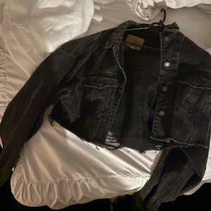Svart croppad jeansjacka - Säljer en svart croppad jeansjacka med råa kanter och knappar framtill. Jackan har en cool, avslappnad stil och är perfekt för att lägga till en edgy touch till din outfit. Den har långa ärmar och klassiska bröstfickor.