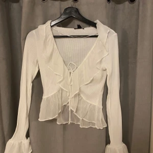 Vit ribbad blus med volangdetaljer - Säljer en stilren vit ribbad blus med volangdetaljer och knyt framtill. Blusen har långa ärmar och en vacker volangkant vid ärmslut. Prislappen sitter kvar så den är i nyskick🫶🏼