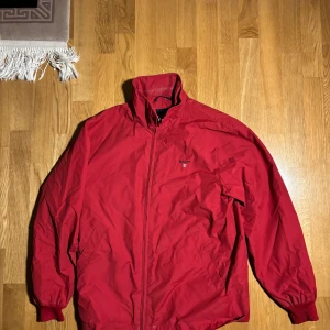 Röd jacka från Gant - Säljer en snygg röd jacka från Gant med dragkedja och broderad logga på bröstet. Jackan har en klassisk design med hög krage och är perfekt för en stilren look.