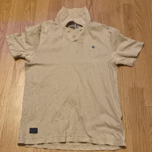 Beige pike från Morris - Säljer en beige kortärmad piké från Morris med en liten blå broderad detalj på bröstet. Piken har en klassisk passform och är tillverkad i ett mjukt material. Perfekt för en avslappnad stil.