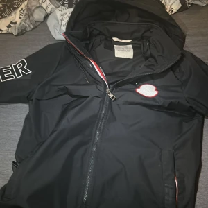 Svart vindjacka från Moncler - Snygg svart vindjacka från Moncler med huva och dragkedja. Jackan har vita och röda detaljer samt Moncler-logga på ärmen. Perfekt för blåsiga dagar och har en stilren design med broderad text på insidan. ÄKTA! Storlek M