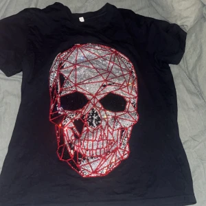 Svart t-shirt med dödskallemotiv - Cool svart t-shirt med ett stort dödskallemotiv i glittrande stenar på framsidan. Motivet har röda och silverfärgade detaljer som ger en edgy look. Perfekt för dig som vill sticka ut! Storlek är M men den sitter som L. Har även storlek S som sitter som M.